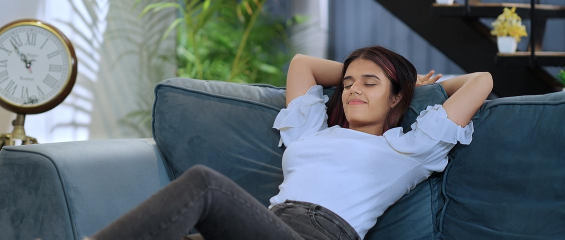 Eine Frau entspannt sich auf einem blauen Sofa mit geschlossenen Augen, trägt ein weißes Top und Jeans, in der Nähe befinden sich eine alte Uhr und Pflanzen.