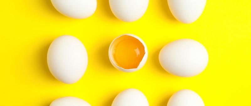 Top view of white, unshelled eggs with one cracked open to reveal orange yolk inside against a vibrant yellow background.