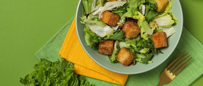 Lettuce and tofu salad in turquoise plate on green and orange napkins beside gold fork and green leafy garnish against a vibrant green background.