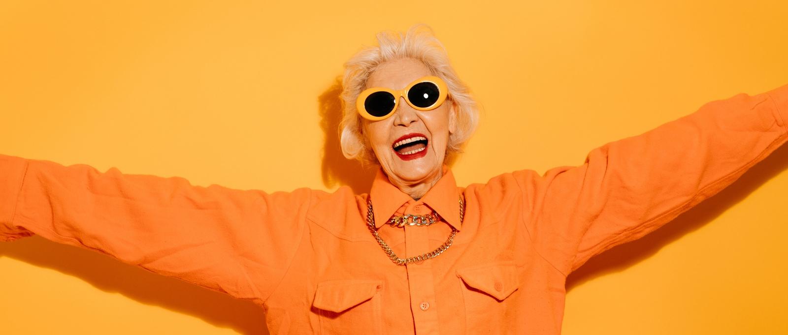 Lächelnde Senior stilvolle Frau trägt trendige Sonnenbrille breitet die Hände, will jemanden zu umarmen, froh, Sie zu sehen isoliert auf gelbem Hintergrund.