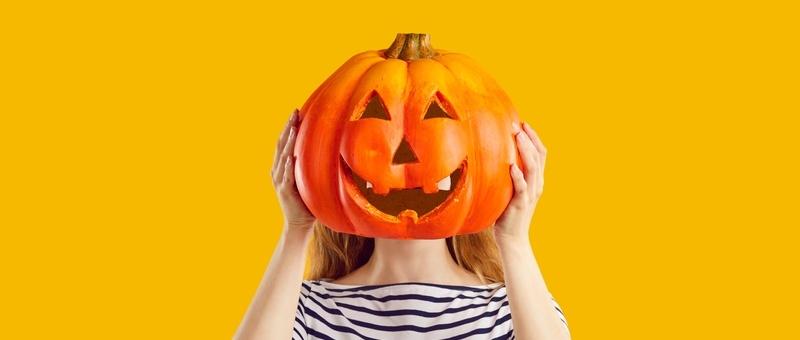 Frau in gestreiftem Oberteil vor gelbem Hintergrund versteckt Gesicht hinter Halloween-Kürbislaterne.