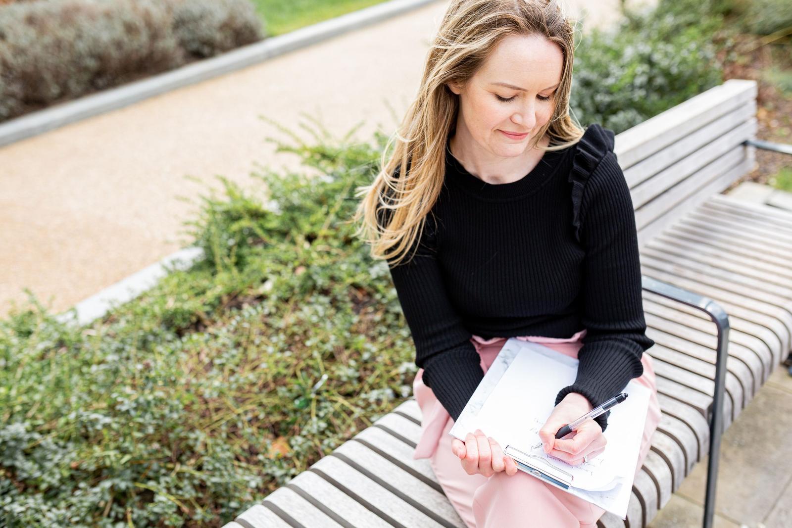 Die anerkannte Psychotherapeutin Laura Greenwood von Laura Greenwood Therapy sitzt mit einem Notizblock auf einer Bank in der Natur.
