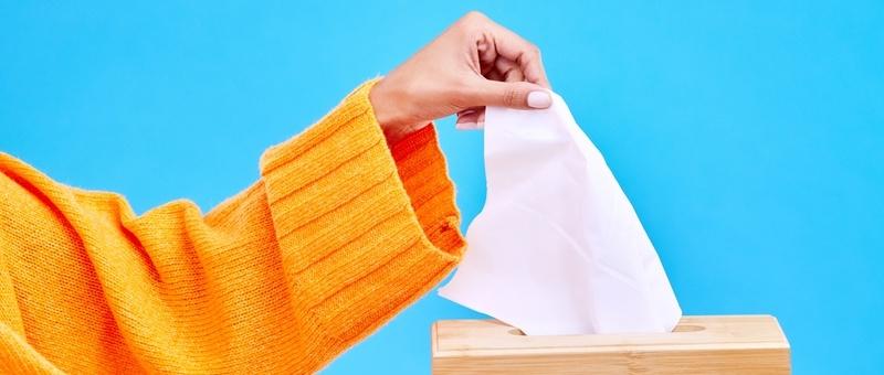 Frau in orangefarbenem Pullover mit Taschentuchbox vor blauem Hintergrund.