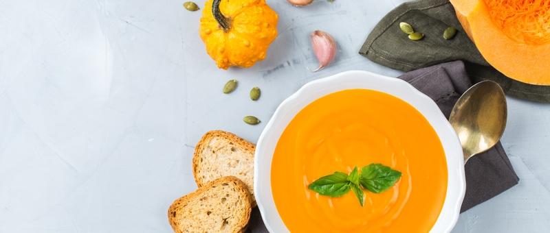 Kürbissuppe in einer Schüssel mit Brotscheiben, Kürbis, Kürbiskernen, Metalllöffel, blauer Serviette und Knoblauchzwiebeln vor einem blassgrauen/blauen Hintergrund.