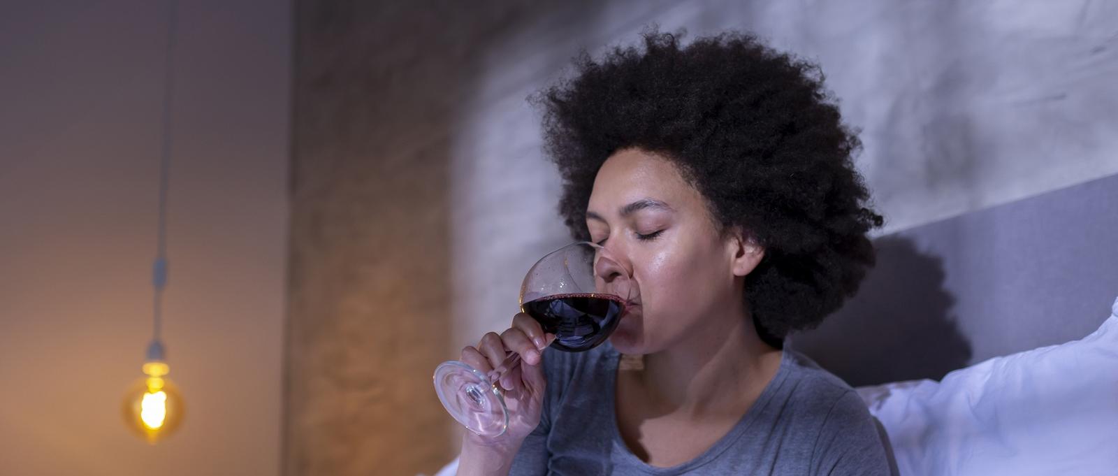 Person mit lockigem Haar, die in einem sanft beleuchteten Raum, in dem eine warme Hängelampe zu sehen ist, Rotwein aus einem Glas schlürft.