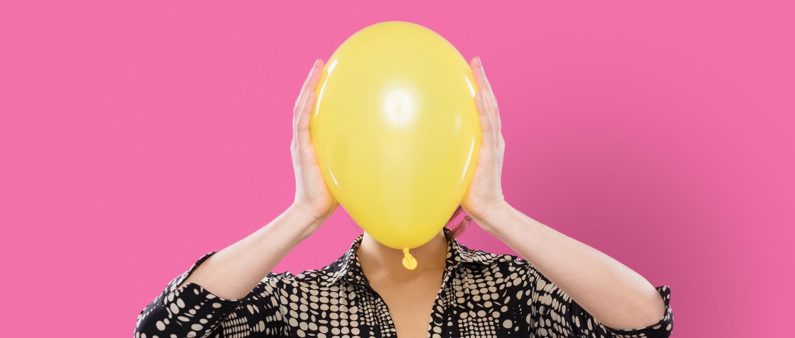 Frau vor rosa Hintergrund, die einen gelben Luftballon über das Gesicht hält.