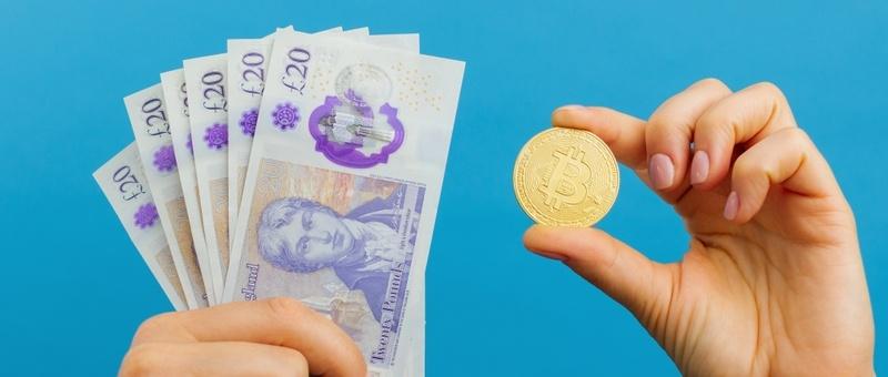 Die Hände halten einen Fächer britischer 20-Pfund-Noten und eine goldene Bitcoin-Münze vor blauem Hintergrund.