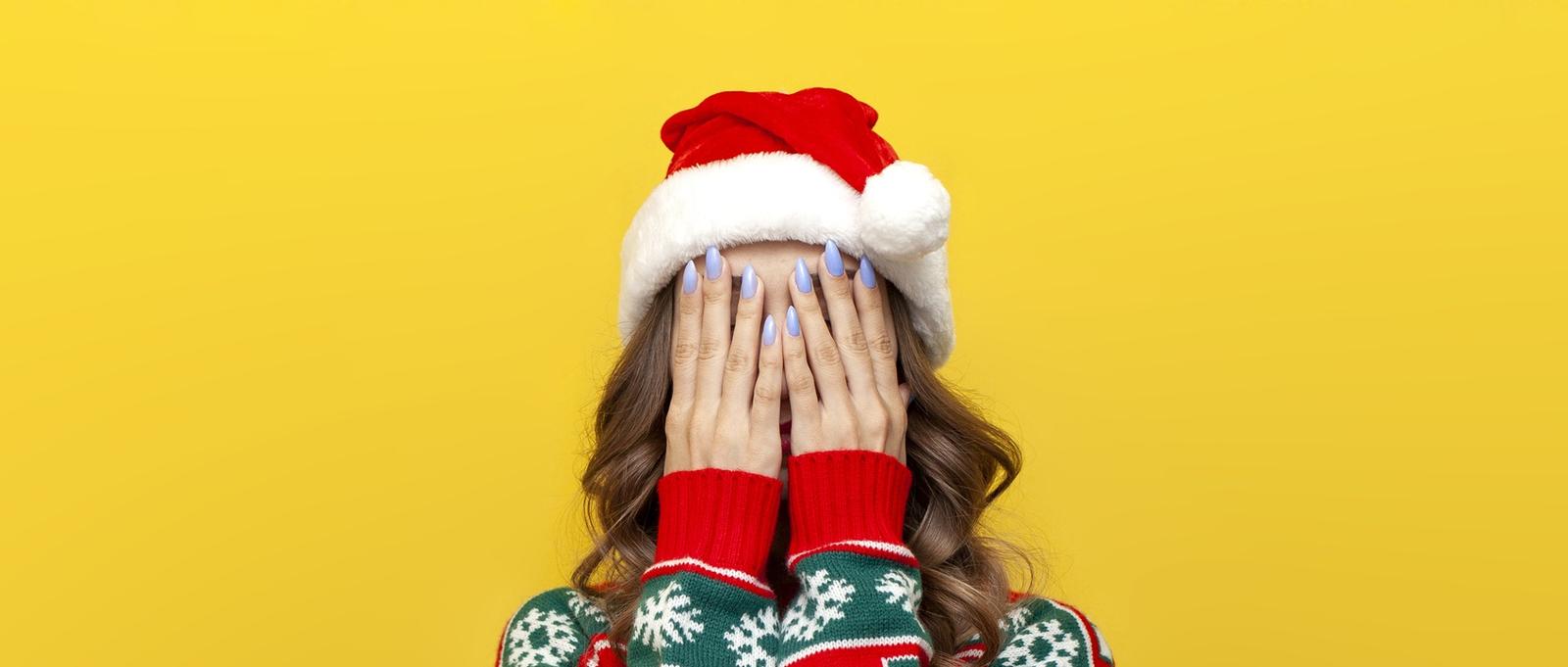 Mädchen in Weihnachtspulli und Weihnachtsmannmütze bedeckt ihr Gesicht mit der Hand und ist schüchtern auf gelbem Hintergrund isoliert, Frau versteckt ihr Gesicht in Stress und Depression in Neujahrskleidung