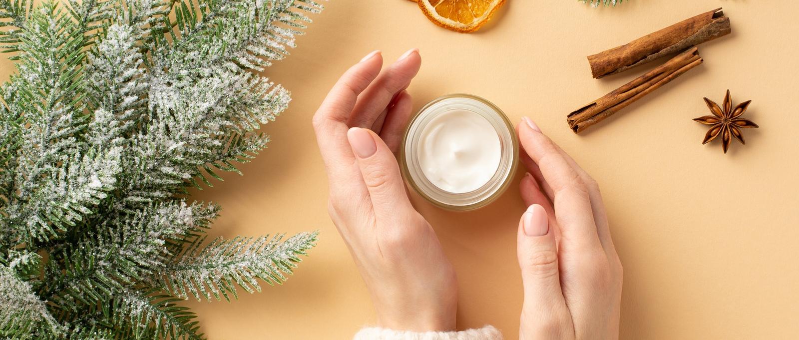 Winter Hautpflege Kosmetik-Konzept. Erste Person Draufsicht Foto von Mädchen Hände in Pullover Cremetopf Fichte Zweige getrocknete Orangenscheiben Zimtstangen und Anis auf isolierten Pastell beige Hintergrund