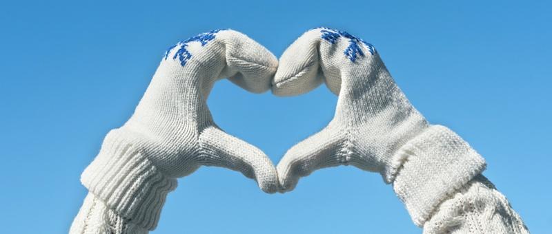 Person, die Herzhände macht, trägt einen gestrickten weißen Pullover und gestrickte weiße Handschuhe mit Schneeflockenaufdrucken vor einem winterlichen blauen Himmel.