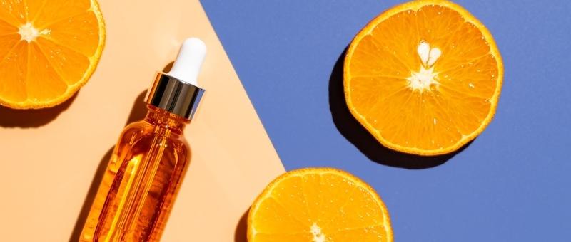 Orangefarbene Serumflasche mit Tropfer neben frischen Orangenhälften auf pfirsichfarbenem und blauem Hintergrund.
