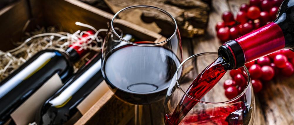 Rotwein, der in ein Glas gegossen wird, neben einem gefüllten Glas Rotwein, mit Flaschen und frischen roten Trauben auf einer Holzfläche.