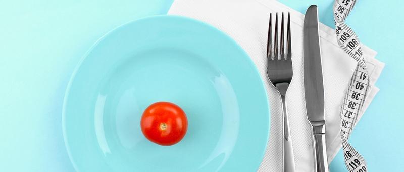 Rote Kirschtomate auf einem weißen Teller neben Maßband und Besteck. Blauer Hintergrund.