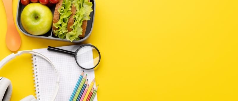 Lunchpaket mit Schulutensilien vor einem gelben Hintergrund.