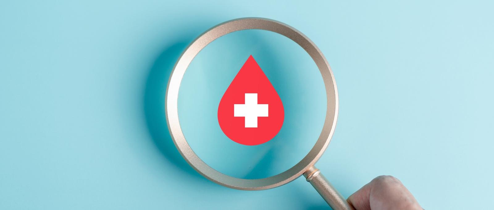 Hand holding magnifying glass focusing on red blood drop icon on pale blue background.
