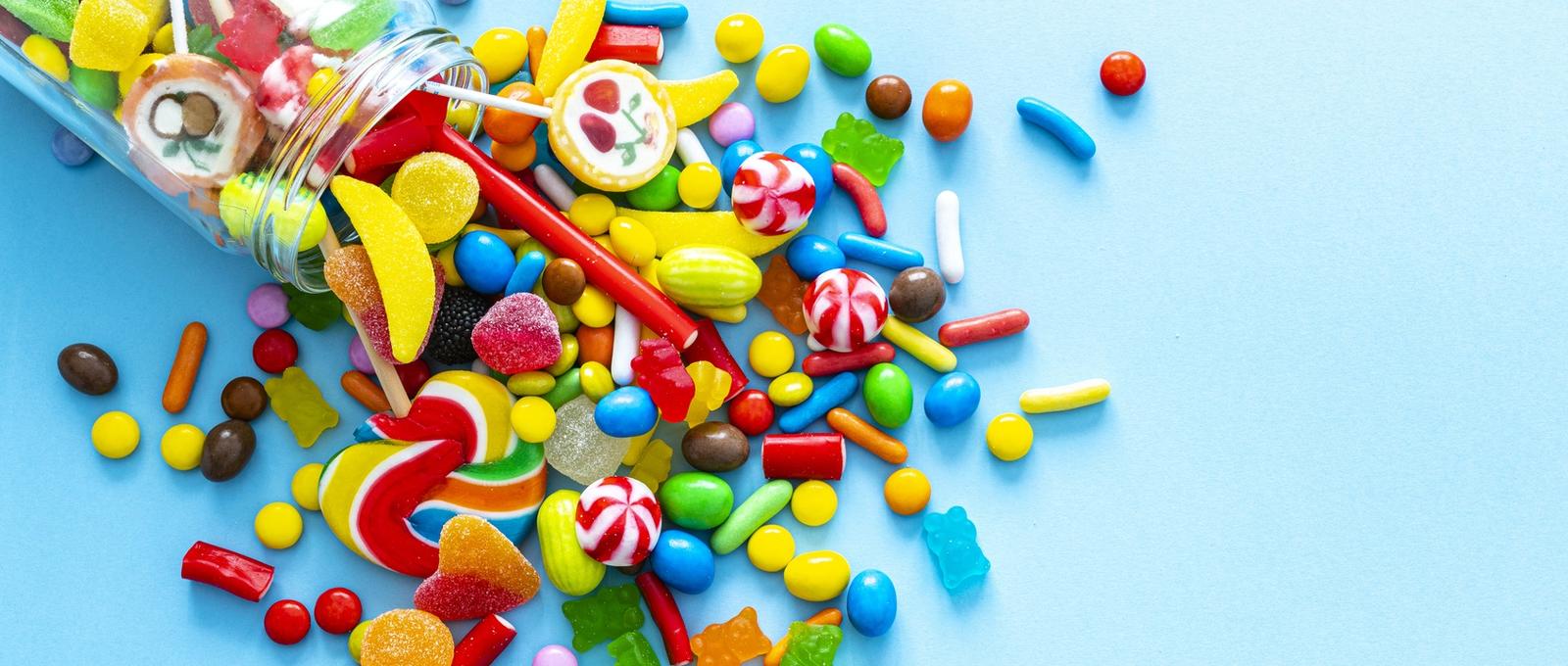 Große Gruppe aller Arten von bunten Bonbons, die aus einem Glasgefäß herausquellen, von oben auf blauem Hintergrund aufgenommen.