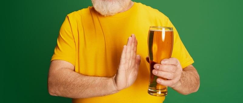 Porträt eines älteren Mannes in gelbem T-Shirt, der mit Bier auf grünem Hintergrund posiert. Alkoholische Getränke ablehnend.