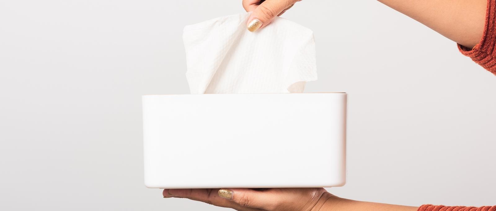 Hand mit goldenem Nagellack, die ein Taschentuch aus einer weißen rechteckigen Schachtel vor einem hellgrauen Hintergrund zieht.