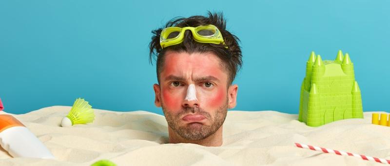 Im Sand vergrabener Mann mit Badespielzeug und sonnenverbranntem Gesicht.