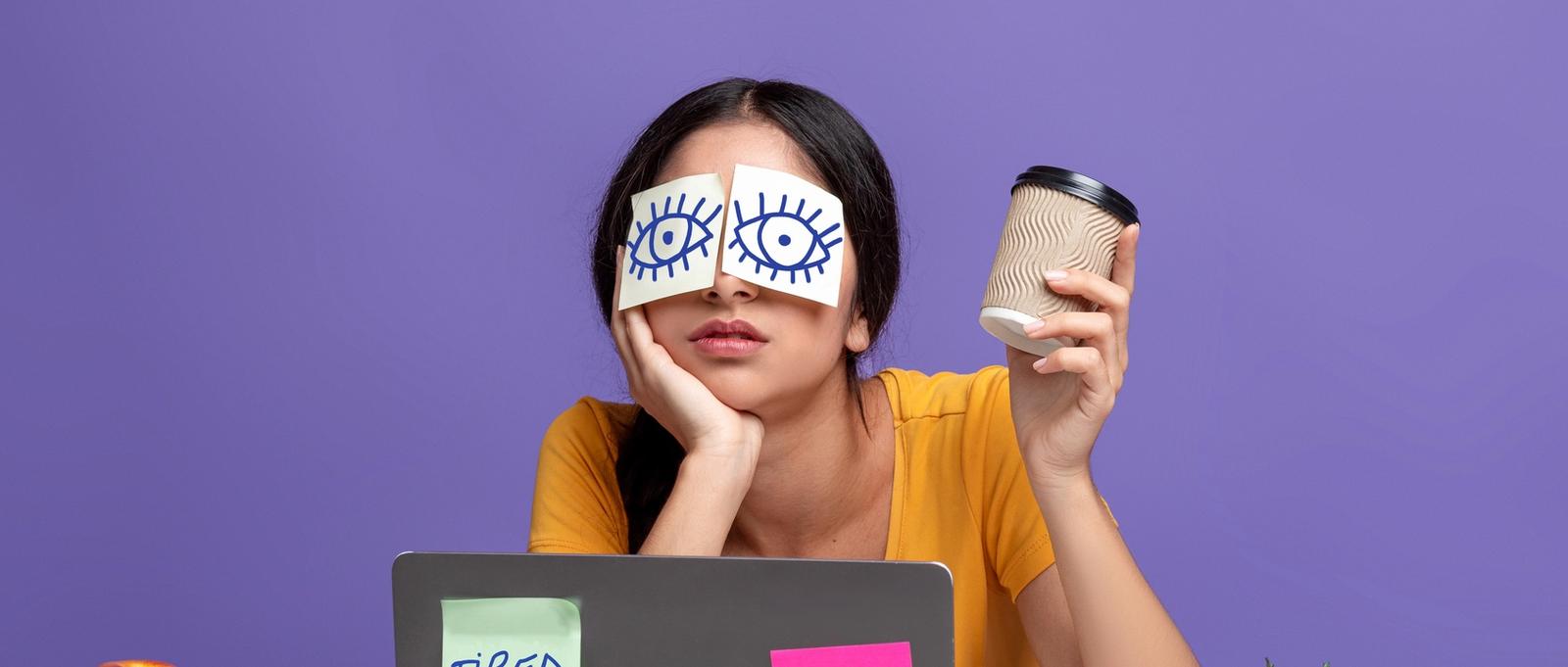 Porträt einer erschöpften jungen Frau, die am Arbeitsplatz sitzt, mit Aufklebern auf den Augen und einer Tasse mit Kaffee in der Hand, die am Laptop am Tisch vor einem lila Studiohintergrund arbeitet