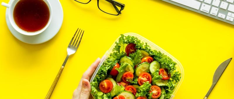 Office scene - pair of male Caucasian hands holding salad bowl on bright yellow table background, surrounded by silver laptop, black rectangular spectacles, silver cutlery, and black tea/coffee in white cup.