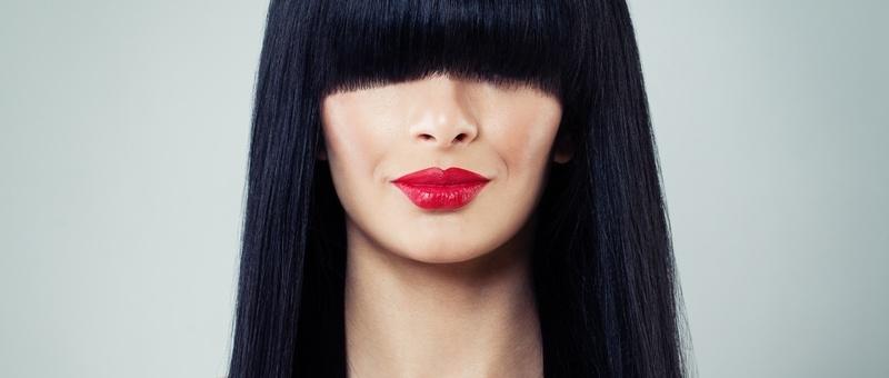 Female wearing dark, red/pink lipstick with dark hair and bangs covering her eyes against a pale grey background.