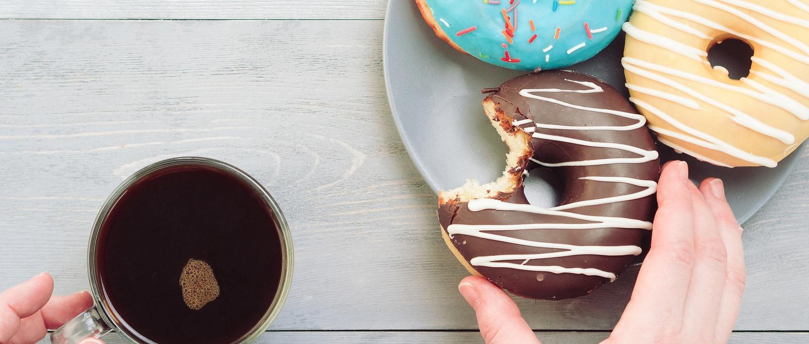Frau Hände mit Kaffee und verschiedene Krapfen über grauen Holztisch. Draufsicht auf bunte Krapfen und Kaffee auf grauem Holzhintergrund.