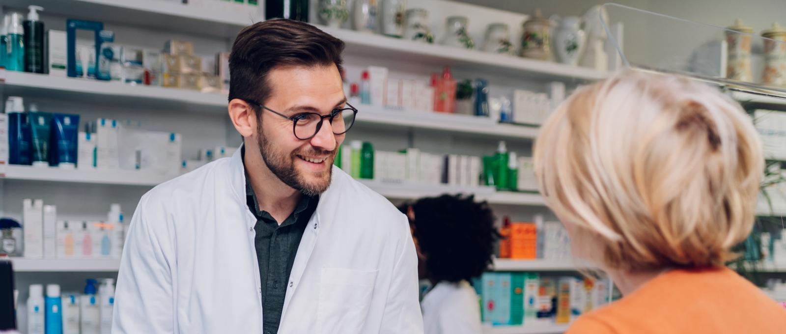 Junger männlicher Apotheker im Gespräch mit einer Kundin in einer Apotheke mit einer Apothekerin im Hintergrund. Regale mit Gesundheitsprodukten.