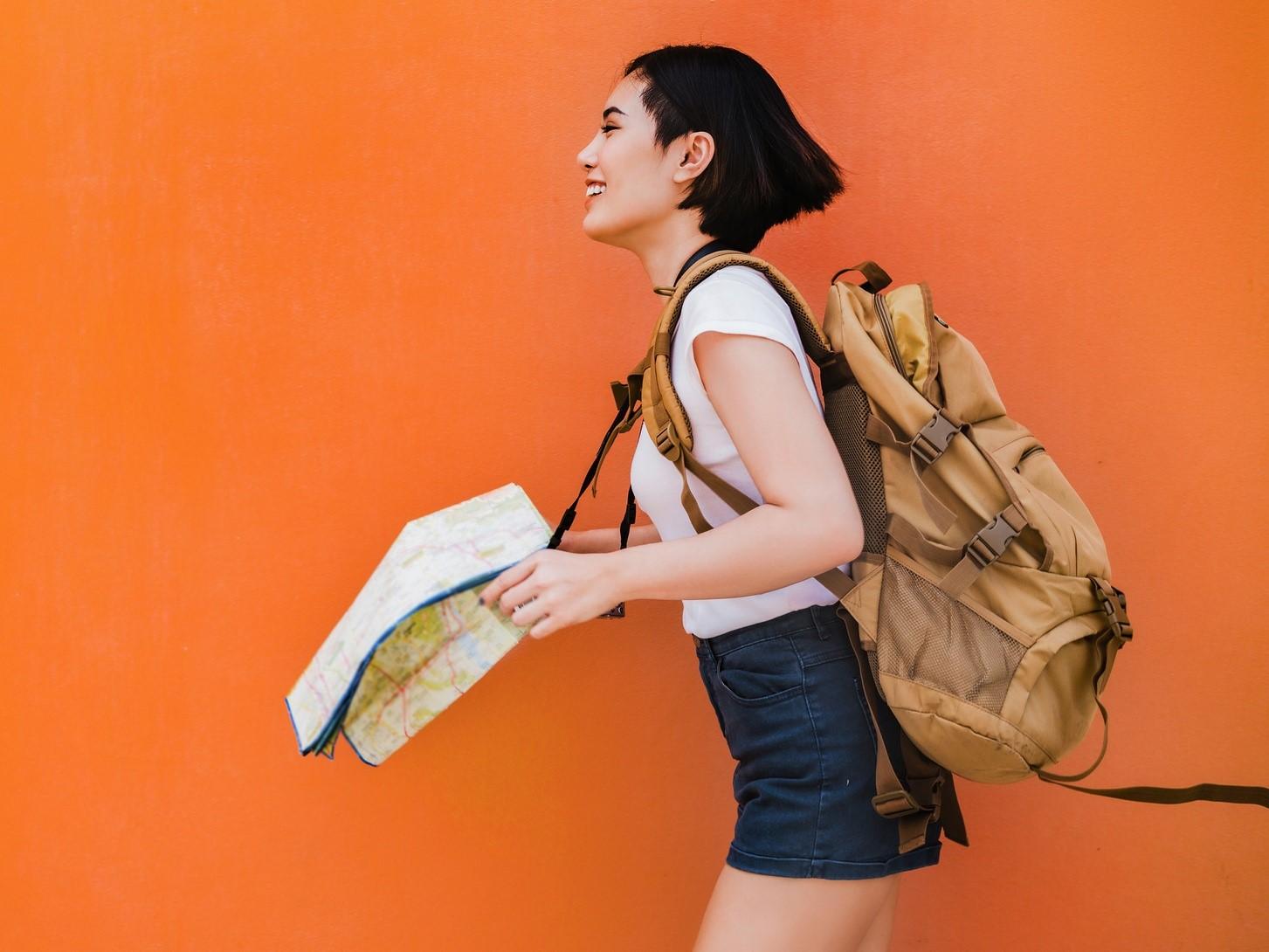 Person mit kurzen dunklen Haaren, die einen Rucksack trägt und eine Landkarte in der Hand hält, lächelnd vor einem leuchtend orangefarbenen Hintergrund.