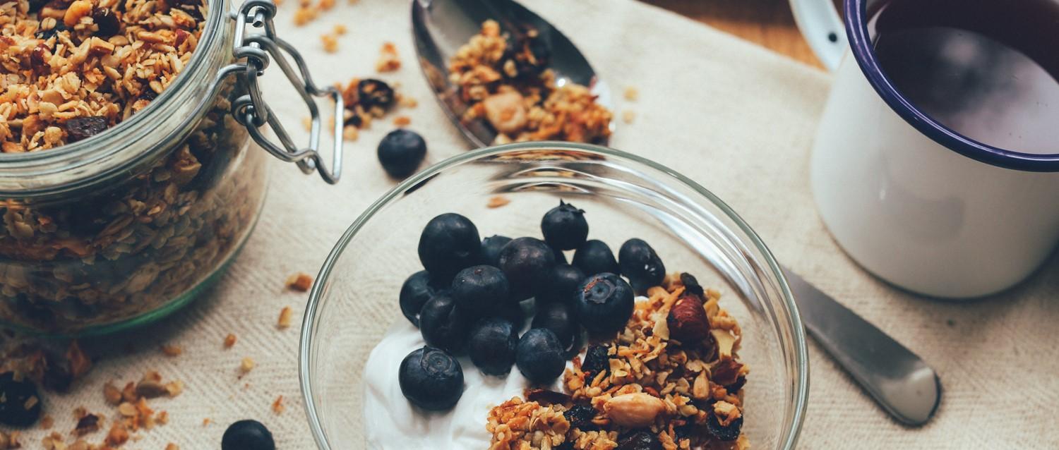Frische Blaubeeren und hausgemachtes Müsli in einer Glasschale mit Joghurt, daneben ein Vorratsglas mit Müsli.