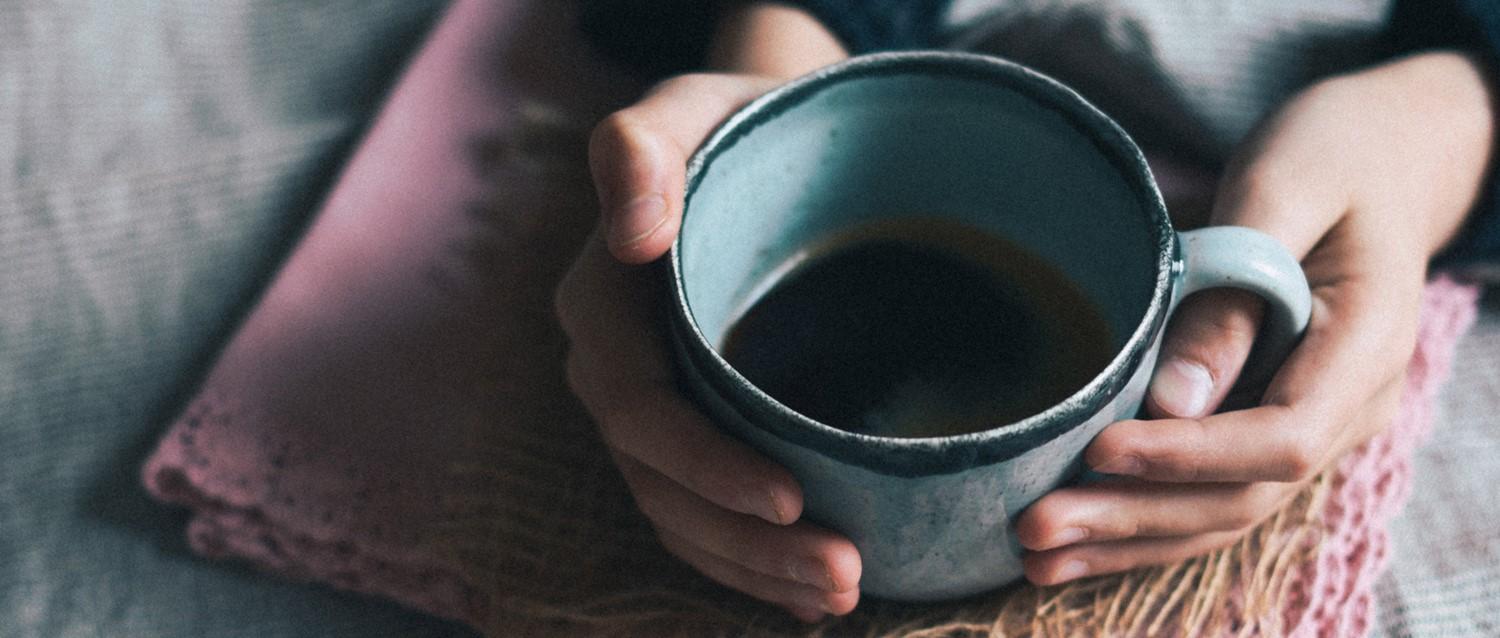 Die Hände halten einen blauen, mit Kaffee gefüllten Keramikbecher, der auf einem rosafarbenen Textil liegt, das eine gemütliche, intime Atmosphäre schafft.