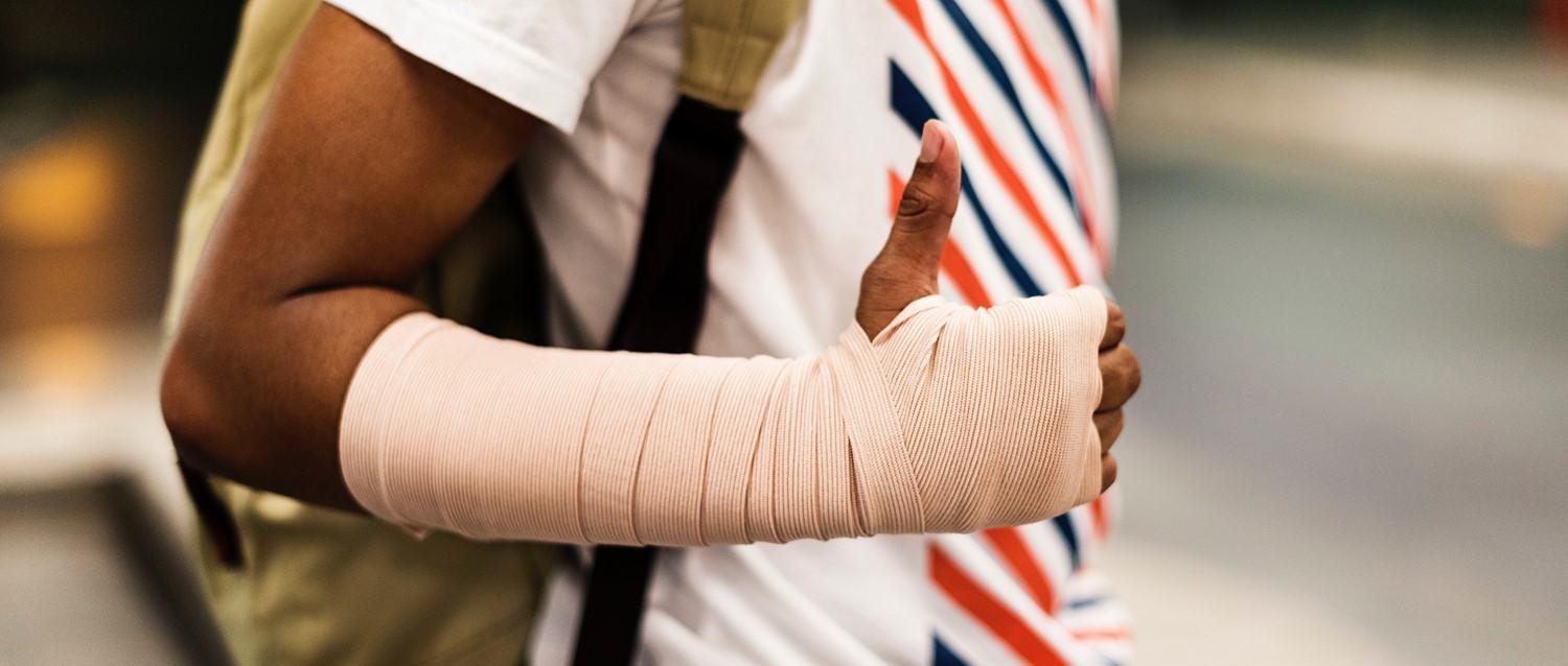 Person, die ein weißes Hemd mit roten und blauen Streifen trägt und eine Hand und einen Unterarm zeigt, die mit einem beigen Verband umwickelt sind.