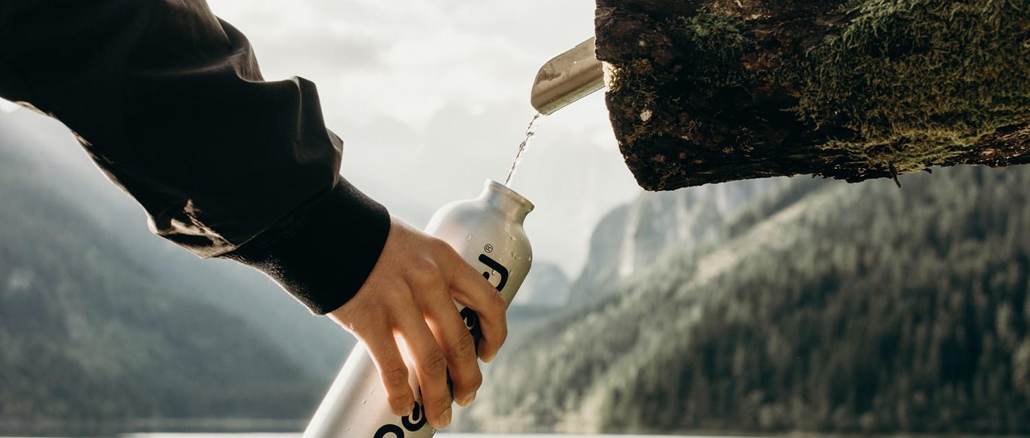 Eine Person füllt eine wiederverwendbare Wasserflasche aus einer natürlichen Quelle in einer Berglandschaft mit dramatischen Felsgipfeln im Hintergrund.