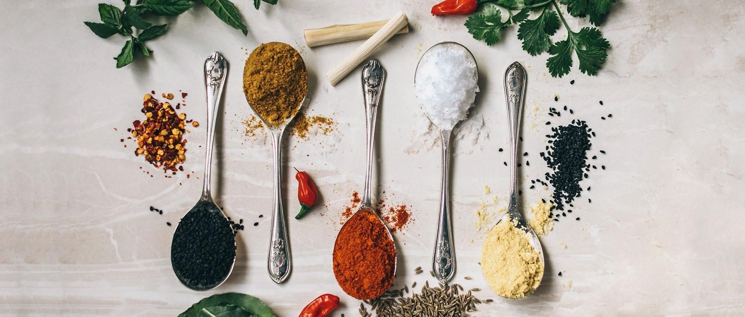 Silberlöffel mit bunten Gewürzen wie Curry, Paprika, Salz und schwarzen Samen, umgeben von frischen Kräutern und Chilischoten.