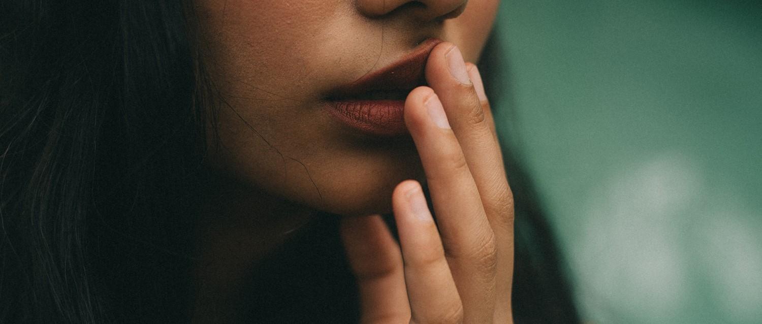 Nahaufnahme einer Person mit roten Lippen, die ihr Gesicht nachdenklich berührt, vor einem sanften grünen Hintergrund.