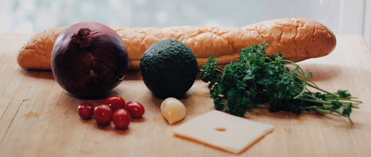 Frische Zutaten auf Holzbrett: Baguette, rote Zwiebel, Avocado, Petersilie, Kirschtomaten, Knoblauch und Käsescheibe.