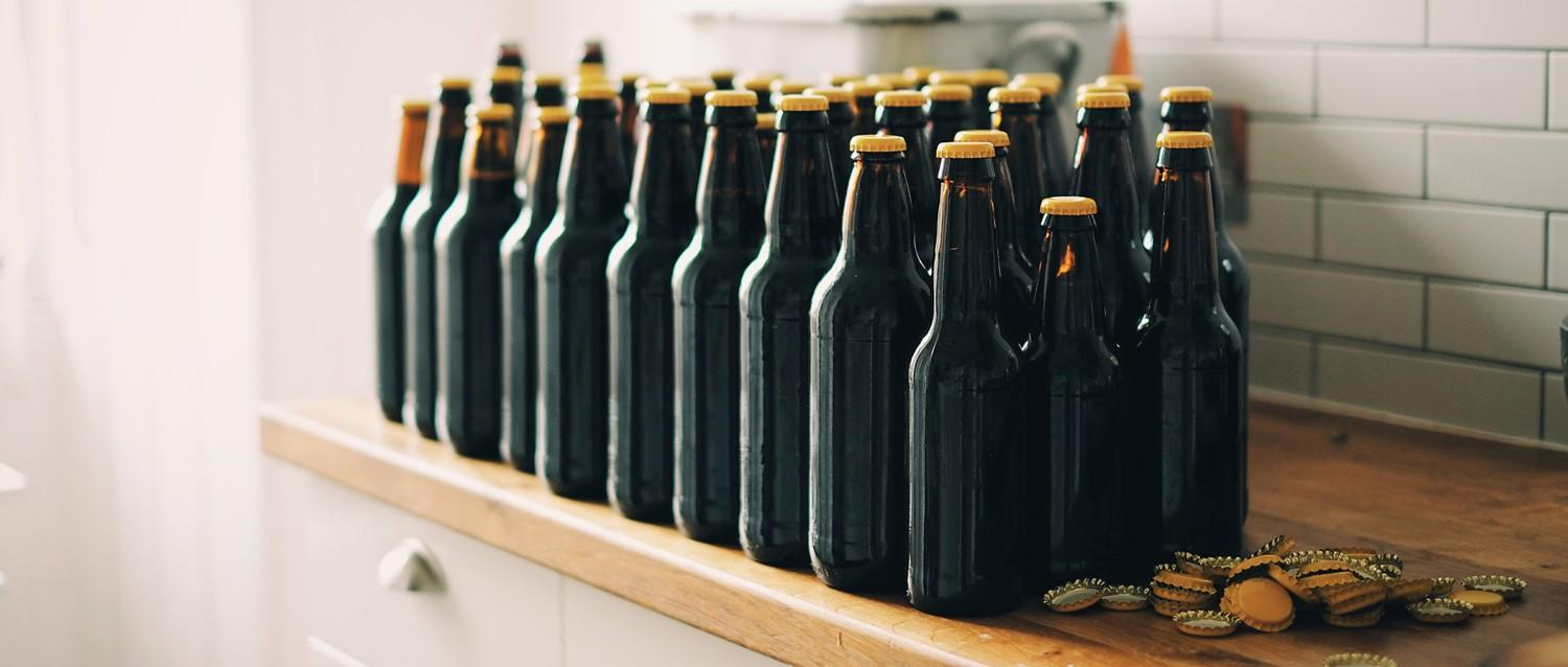 Auf einer hölzernen Arbeitsplatte stehen reihenweise Bierflaschen aus dunklem Glas mit goldenen Verschlüssen, und in der Nähe liegen verstreut Flaschenverschlüsse.