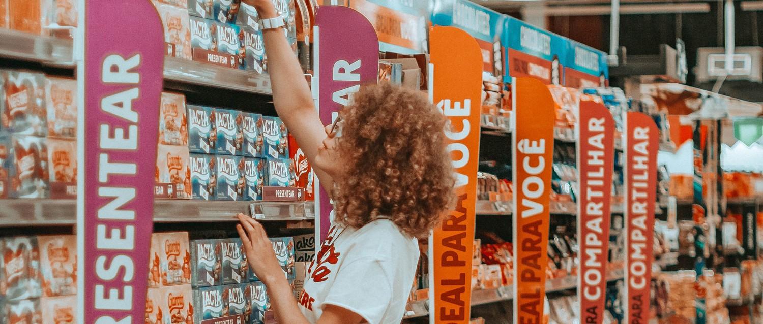 Person mit lockigem Haar, die in einem Supermarktgang mit bunten Kategorienschildern in Pink und Orange nach Produkten greift.