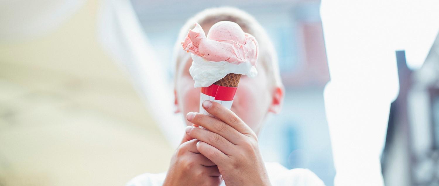 Hände halten eine Erdbeereiswaffel mit rosa Kugeln und Schlagsahne vor einem hellen, unscharfen Hintergrund.
