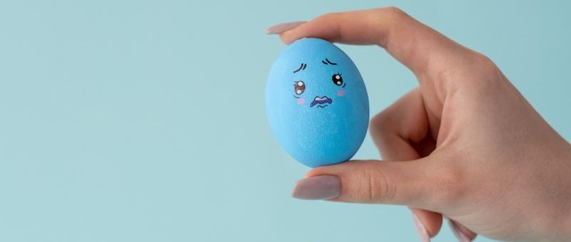 Eine Hand hält ein trauriges, blaues, hart gekochtes Ei vor blauem Hintergrund.