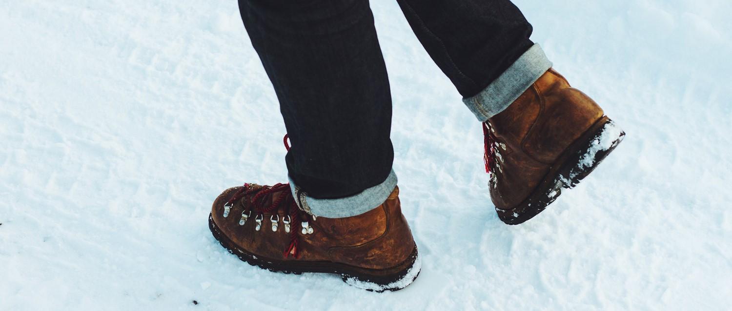 Person in braunen Wanderstiefeln mit roten Schnürsenkeln und hochgekrempelten dunklen Jeans geht durch den Schnee.