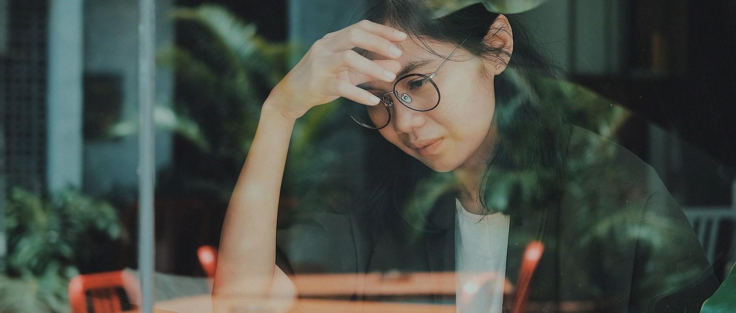 Person mit Brille, gestresst wirkend, Hand auf der Stirn, am Fenster sitzend, mit Pflanzenreflexionen in gedämpften Grüntönen.