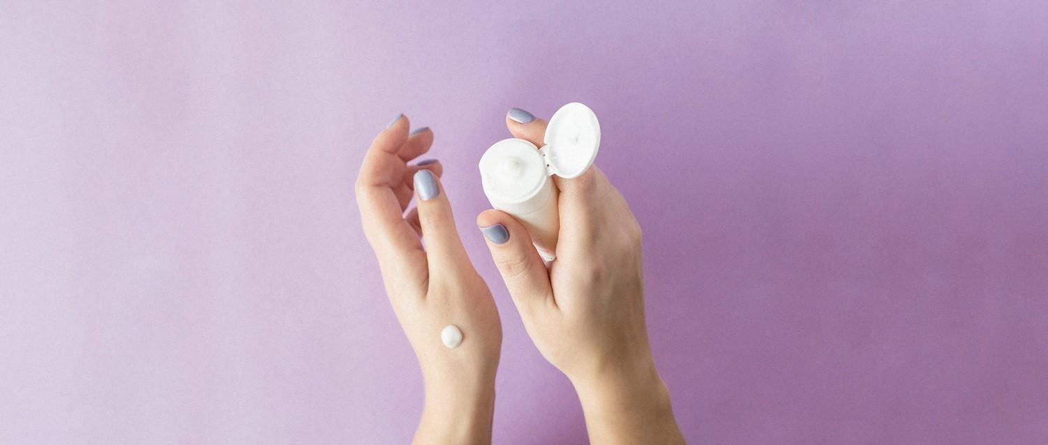 Hände mit blassblauem Nagellack, die einen weißen Hautpflegebehälter mit Creme auf einer Hand vor einem lila Hintergrund halten.
