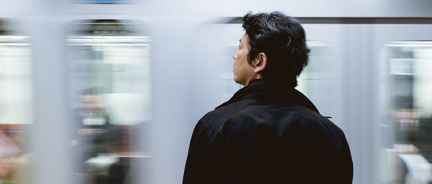 Eine Person in einem schwarzen Mantel wartet auf einem U-Bahn-Bahnsteig, während im Hintergrund ein unscharfer Zug vorbeifährt.