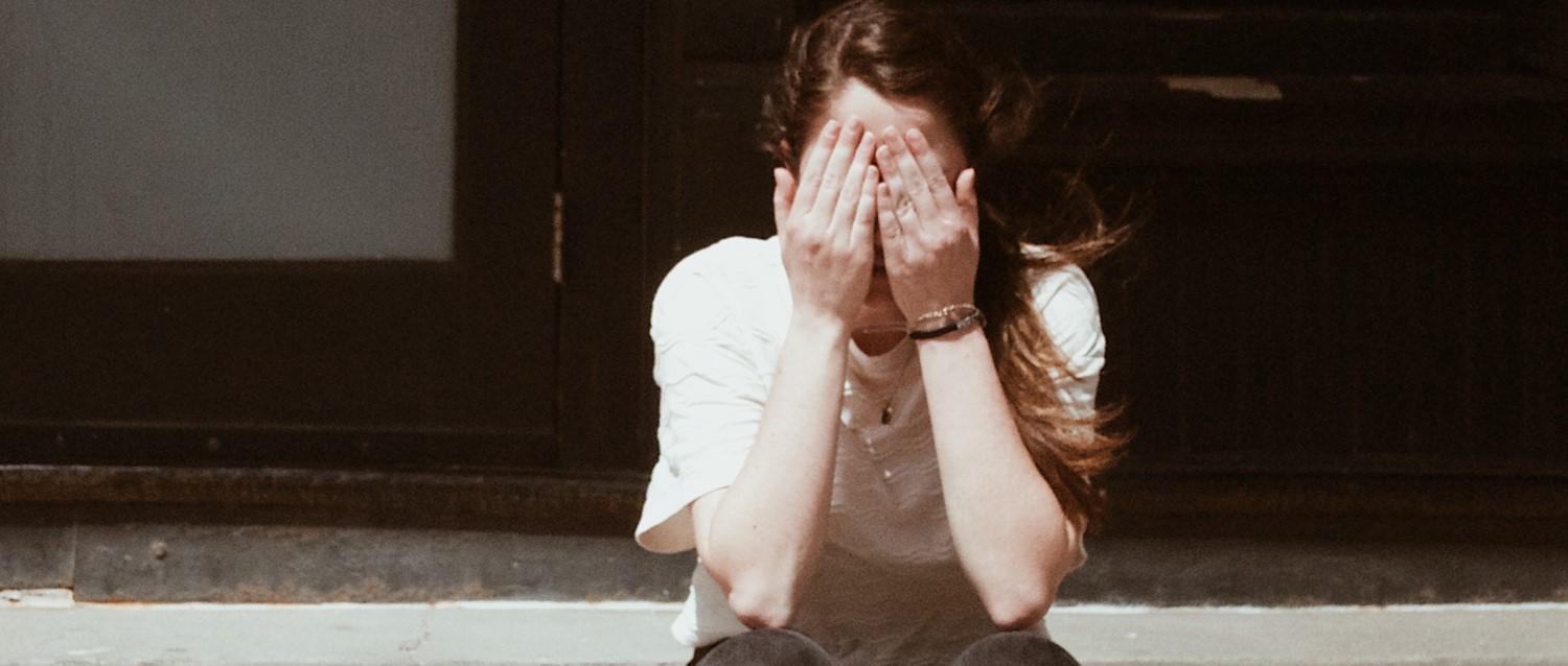 Person in weißem Hemd, die das Gesicht mit den Händen bedeckt und eine Armbanduhr trägt, sitzt vor einem dunklen Hintergrund in einem Bild mit Vintage-Tönen.