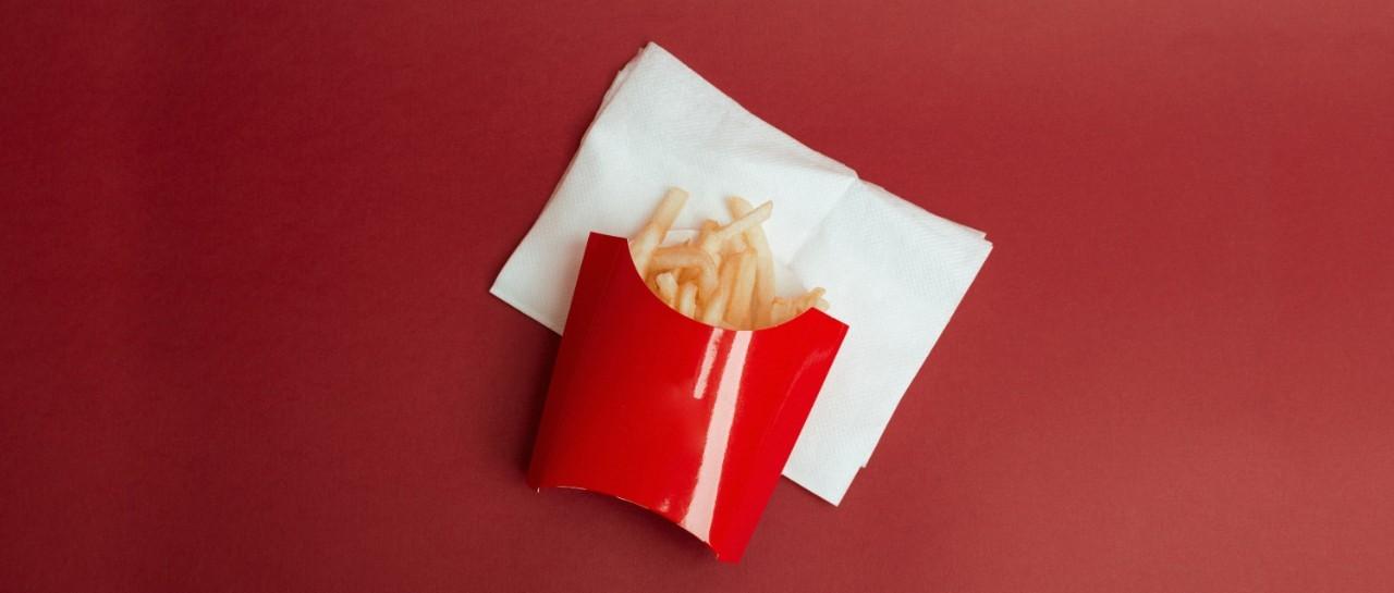 Pommes frites in einer roten Pappschachtel auf einer weißen Serviette vor einem roten Hintergrund.