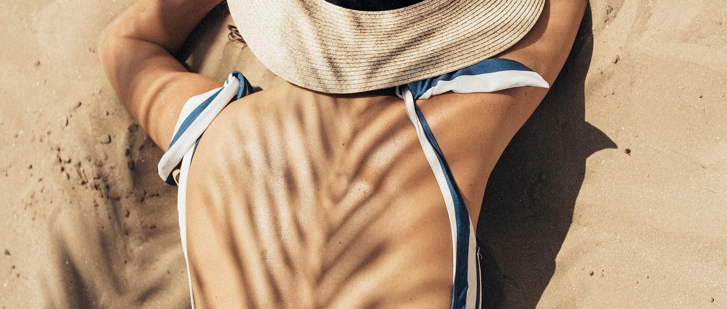 Person, die sich am Sandstrand entspannt, mit Strohhut und blau-weißem Bikini, mit Palmenschatten über ihrem Körper.
