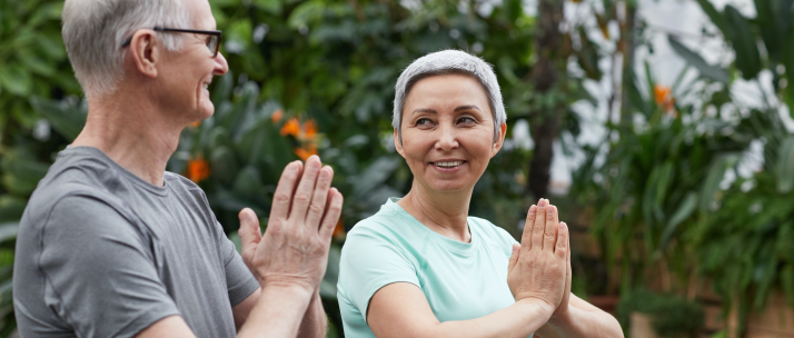 Senior couple practising yoga outdoors, holding prayer pose with hands together against lush green background.
